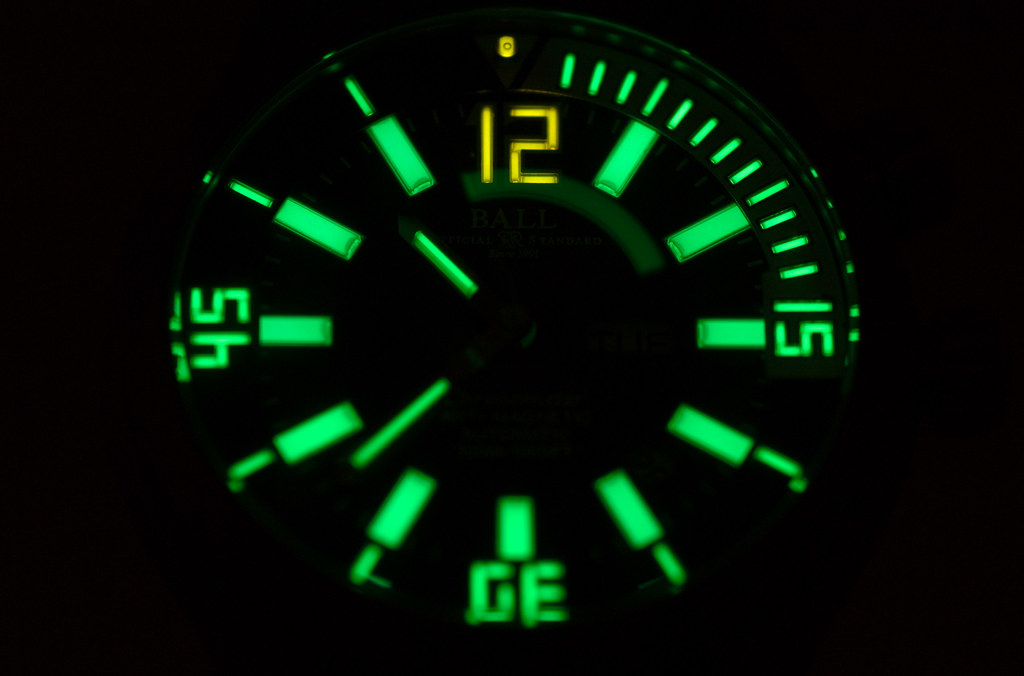 A Ball diver's dial photographed in darkness. Every hour marker, both hands and the minute track of the bezel glow a saturated green; the 12 at the top glows yellow; the 3, 6 and 9 are formed from segmented green tubes. The case, strap and printed text are almost invisible.