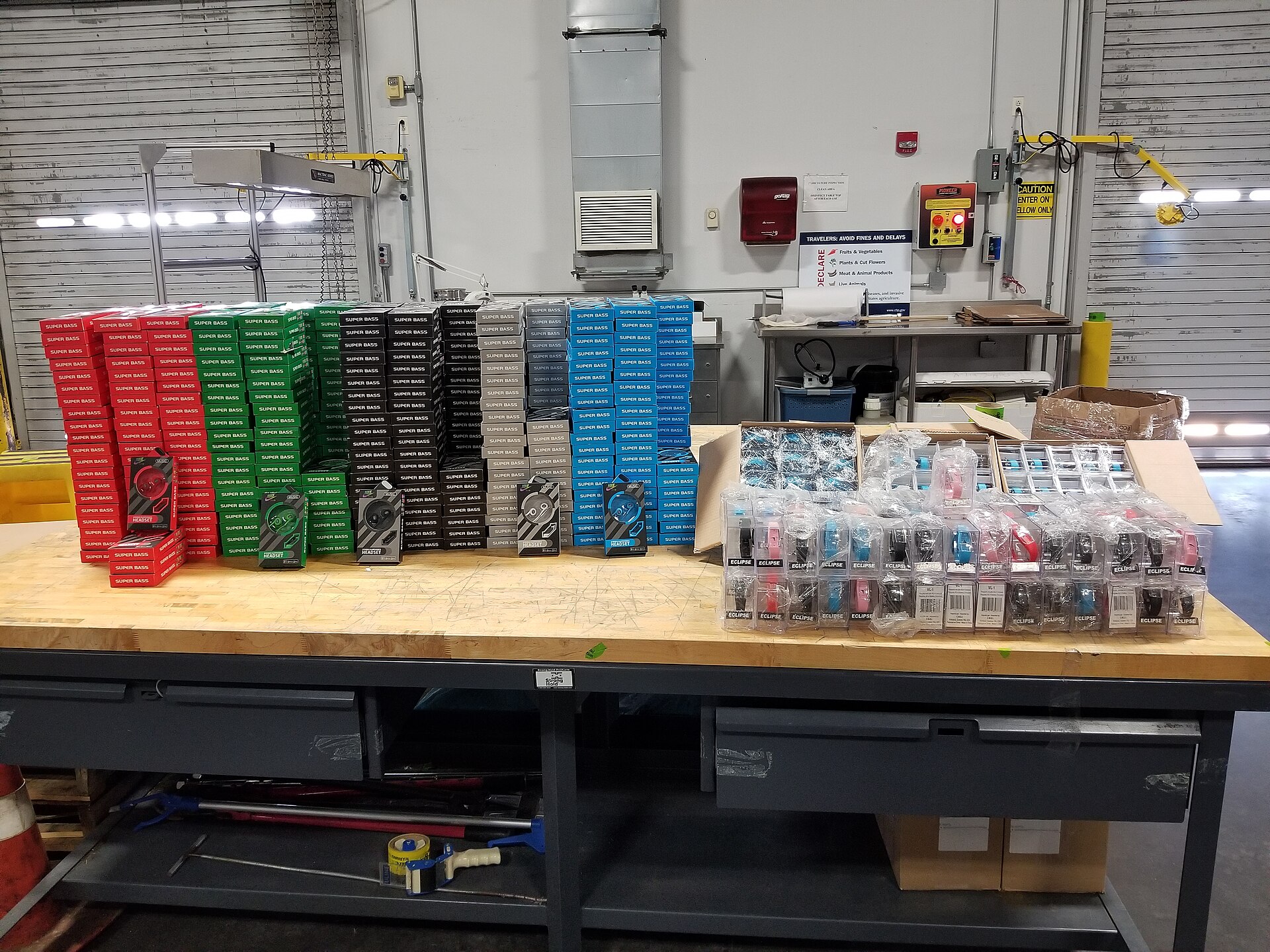 A US Customs and Border Protection officer's gloved hands holding two seized counterfeit wristwatches still in their retail packaging, part of a larger seizure laid out on a table at the Champlain, New York port of entry.