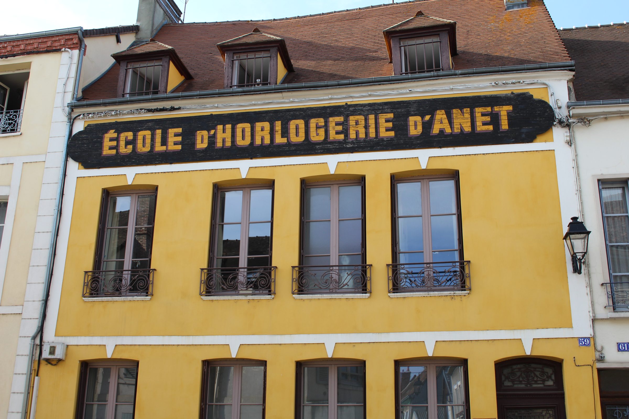 A yellow-painted building facade in Dreux, France, with three dormer windows set into a mansard roof above it. A large black wooden sign spanning the second-floor windows reads "ECOLE D'HORLOGERIE D'ANET" in gilt lettering. Wrought-iron balconies front four tall windows below the sign, and house numbers 59 and 61 are visible on neighbouring doorways.