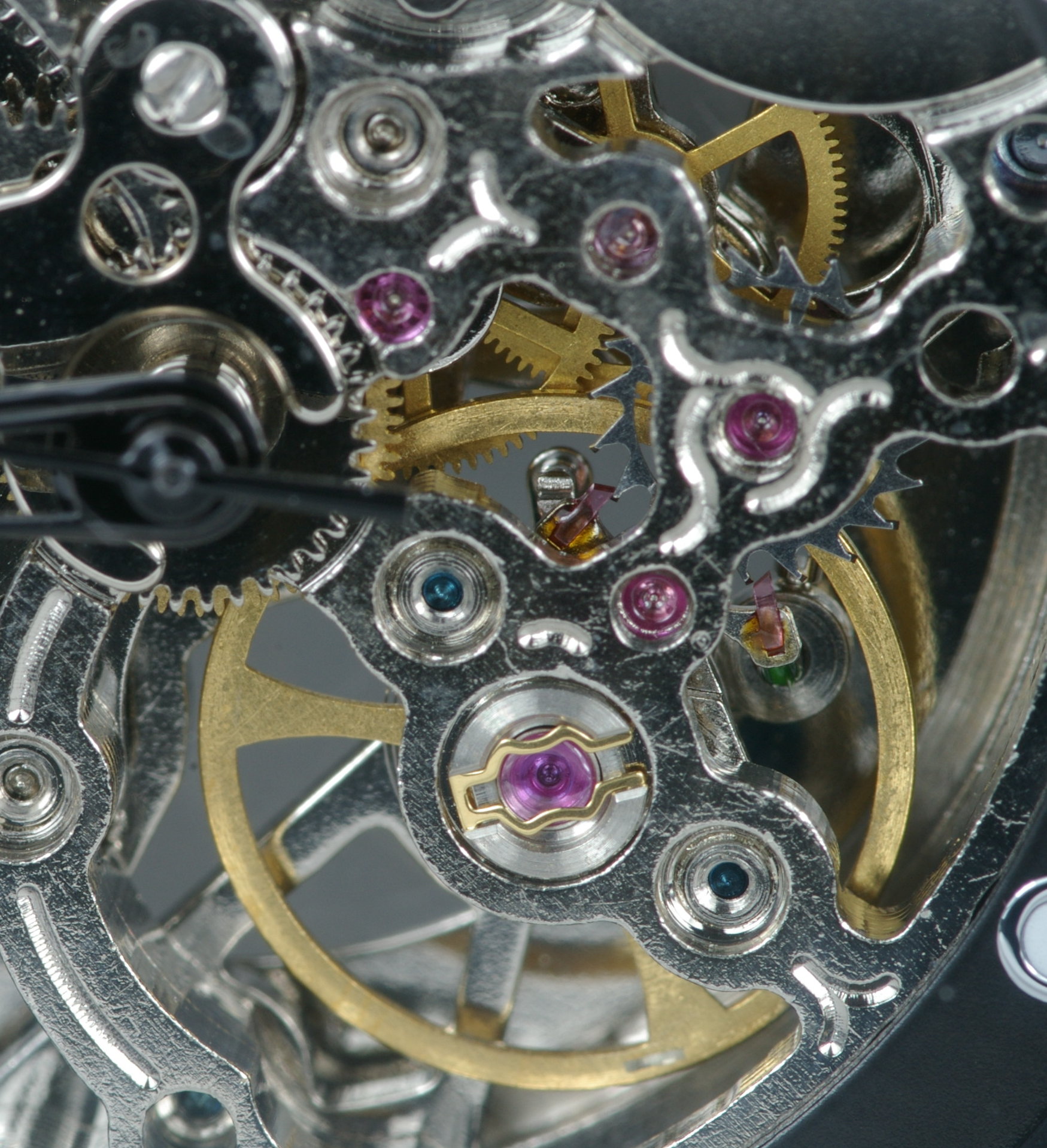 A macro photograph of a skeletonised mechanical watch movement showing the balance wheel, the pallet fork and the escape wheel, with several ruby jewels set into the plate around them.