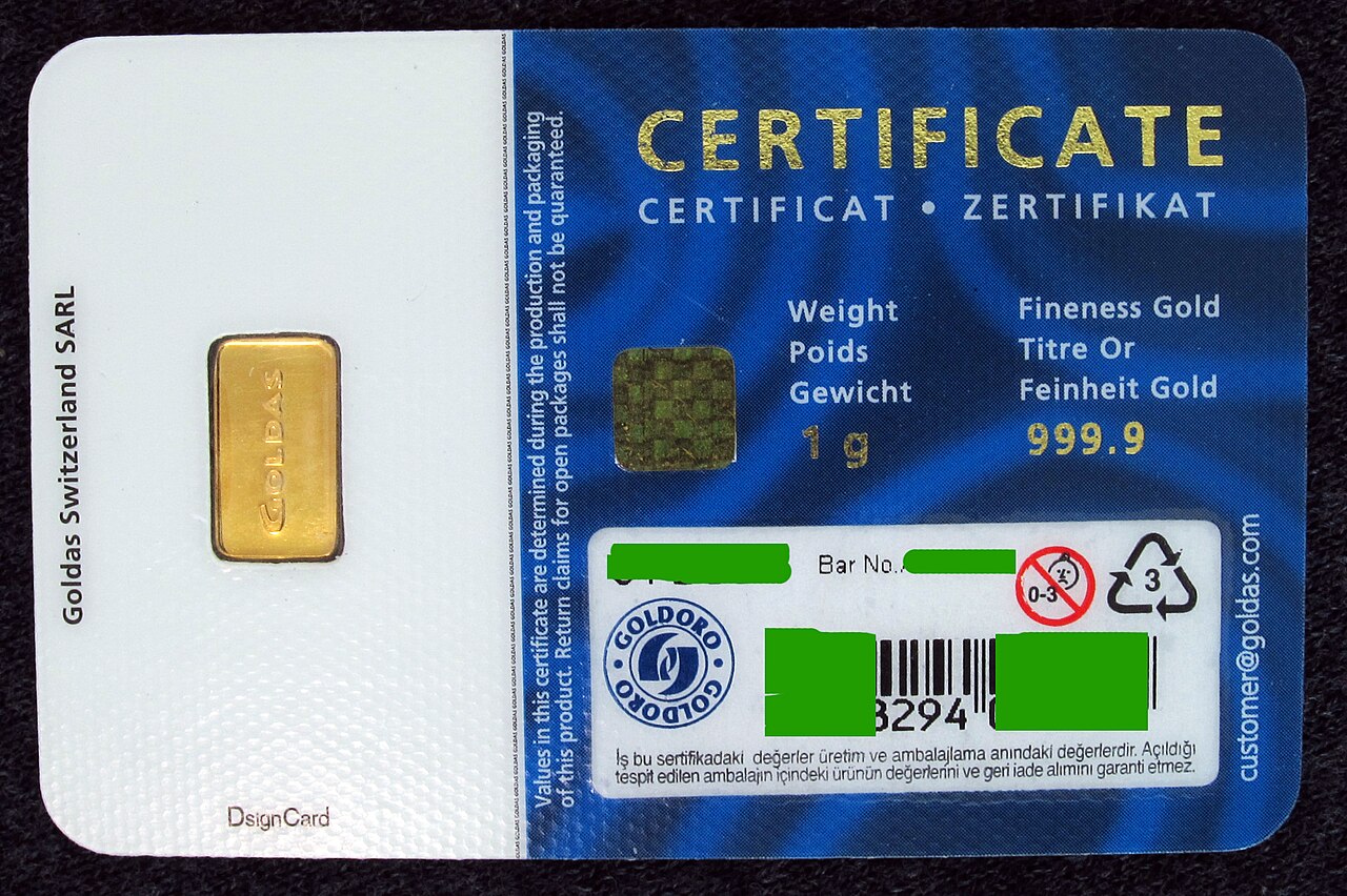 A one-gram gold bar sealed in a Goldas assay card, the card stating a weight of one gram and a fineness of 999.9, with an assay certificate printed alongside.