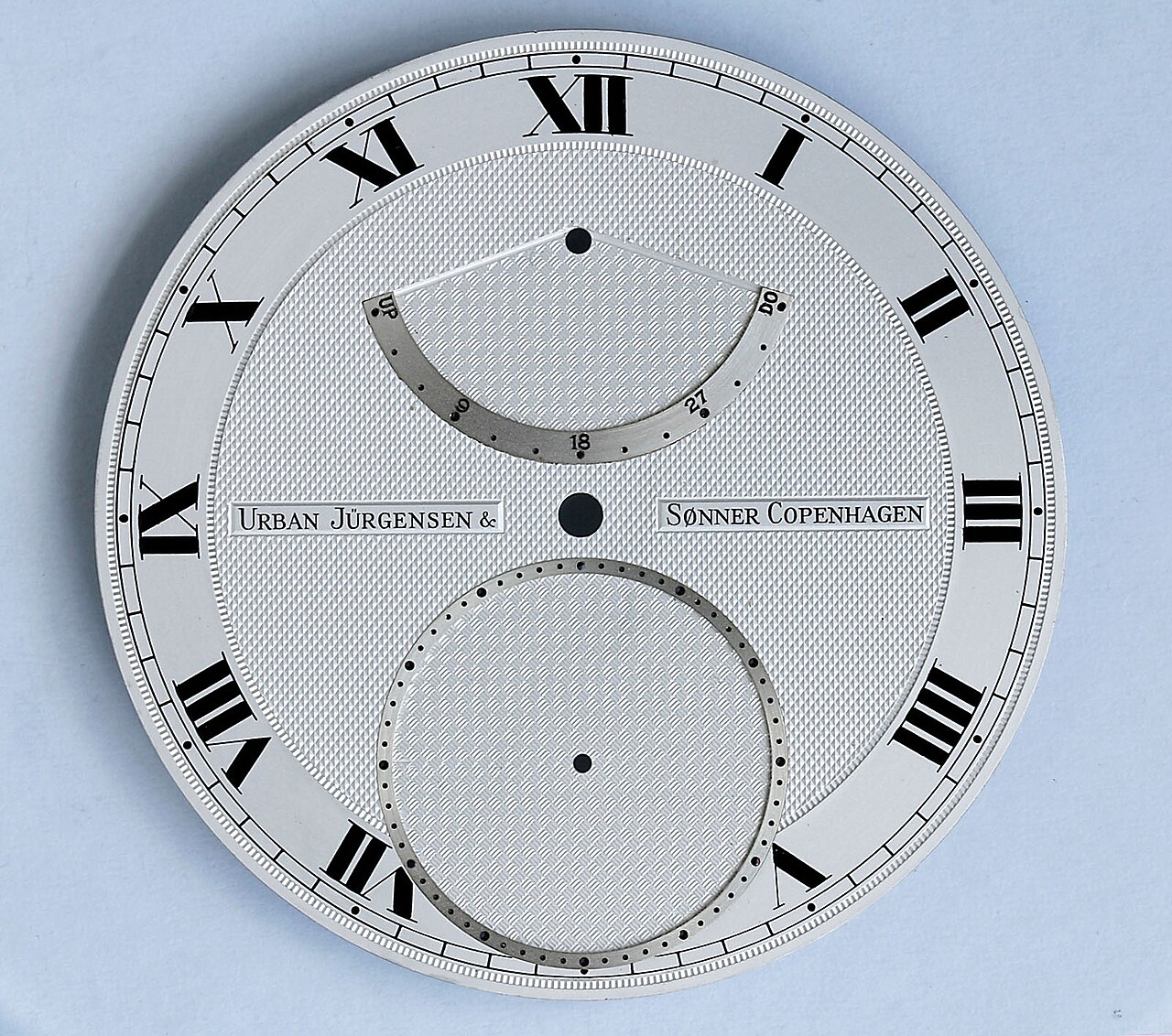 An engine-turned silver watch dial with Roman numerals, a power-reserve sector and a subsidiary seconds register, signed Urban Jürgensen & Sønner Copenhagen.