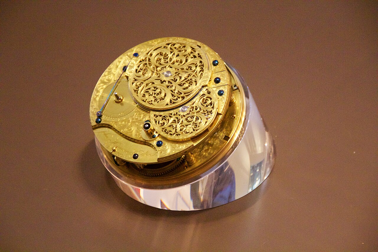 The movement of John Harrison's H4 sea watch, seen from the back out of its case and resting on a clear acrylic stand under museum lighting. The gilt plates are engraved edge to edge with scrollwork; a pierced, engraved balance cock covers most of the upper half, a smaller pierced cock sits below it, and a row of blued steel screws and jewel settings runs around the edge. Harrison's signature is engraved on a cartouche at the lower left.