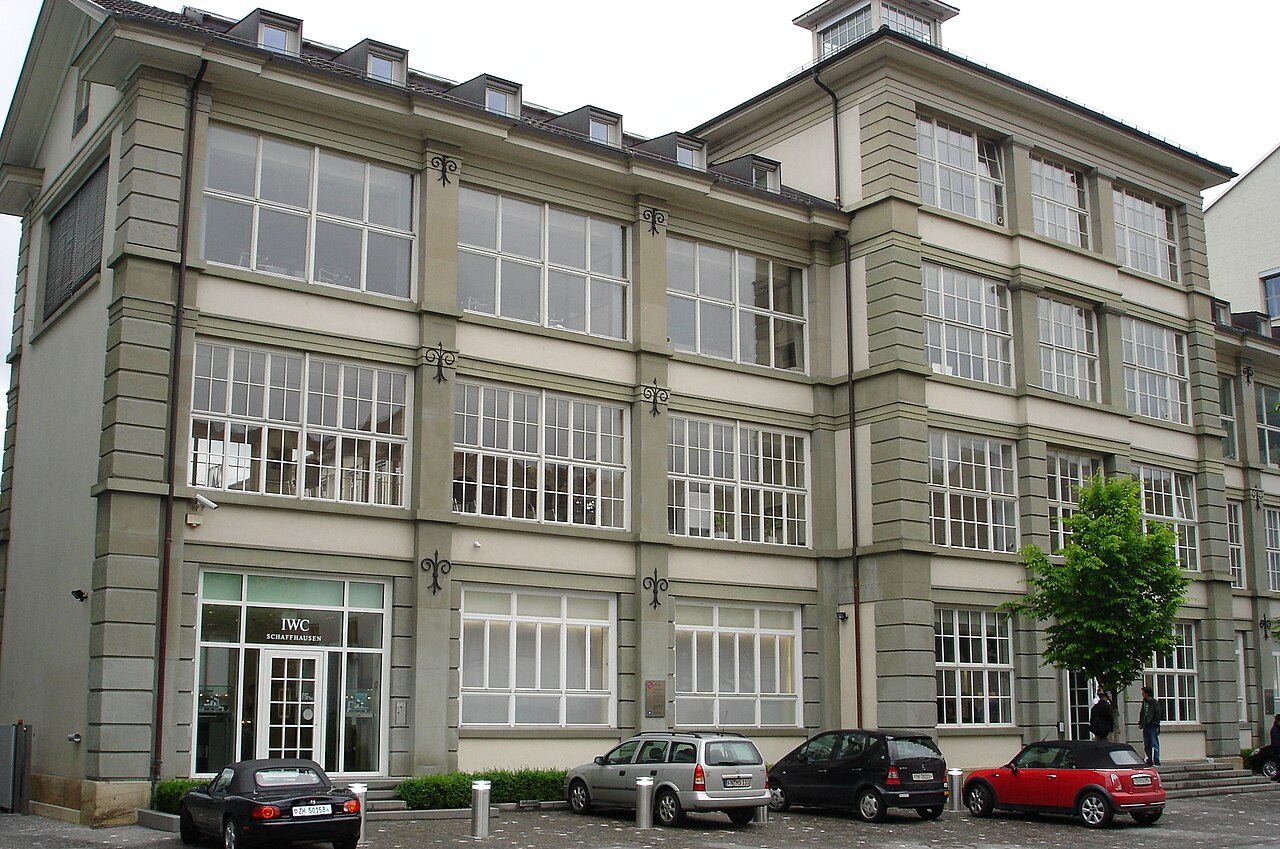 The main entrance of the International Watch Company manufacture in Schaffhausen: a plain early twentieth-century industrial building in pale render, three storeys of tall workshop windows, cars parked along its front.