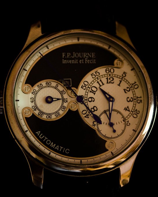 An F.P. Journe Octa in a gold case, lit low against black. The dial carries an off-centre hours and minutes subdial, a power-reserve sector to the left, small seconds below, blued hands, and the signature reading Invenit et Fecit.
