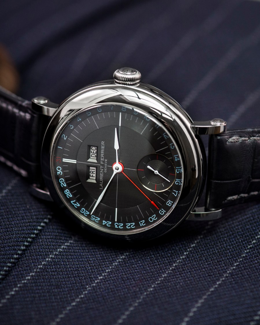 A Laurent Ferrier Annual Calendar in a polished round case with teardrop lugs, photographed at three-quarter angle on navy pinstripe cloth. The slate dial carries day and month apertures, a date track in blue, small seconds and a red central hand.