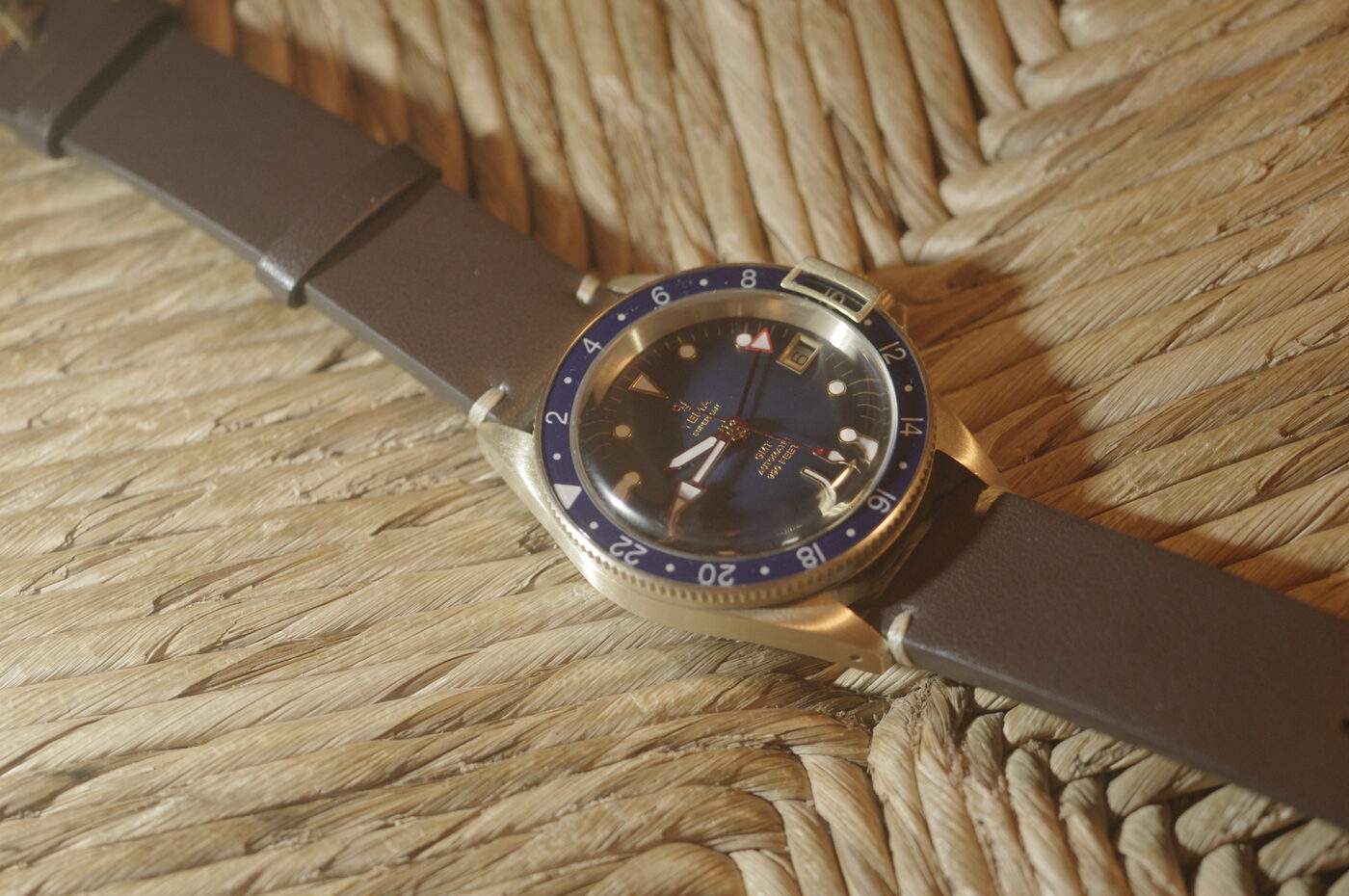 A bronze-cased Yema Superman lying at an angle on a woven straw mat, on a brown leather strap, in warm low light. The bezel is deep blue with a 24-hour scale, and a hinged metal lever stands proud of its edge — the lock that stops the bezel turning. The dial is blue with gilt markers, a date window and a slim red-tipped hand.