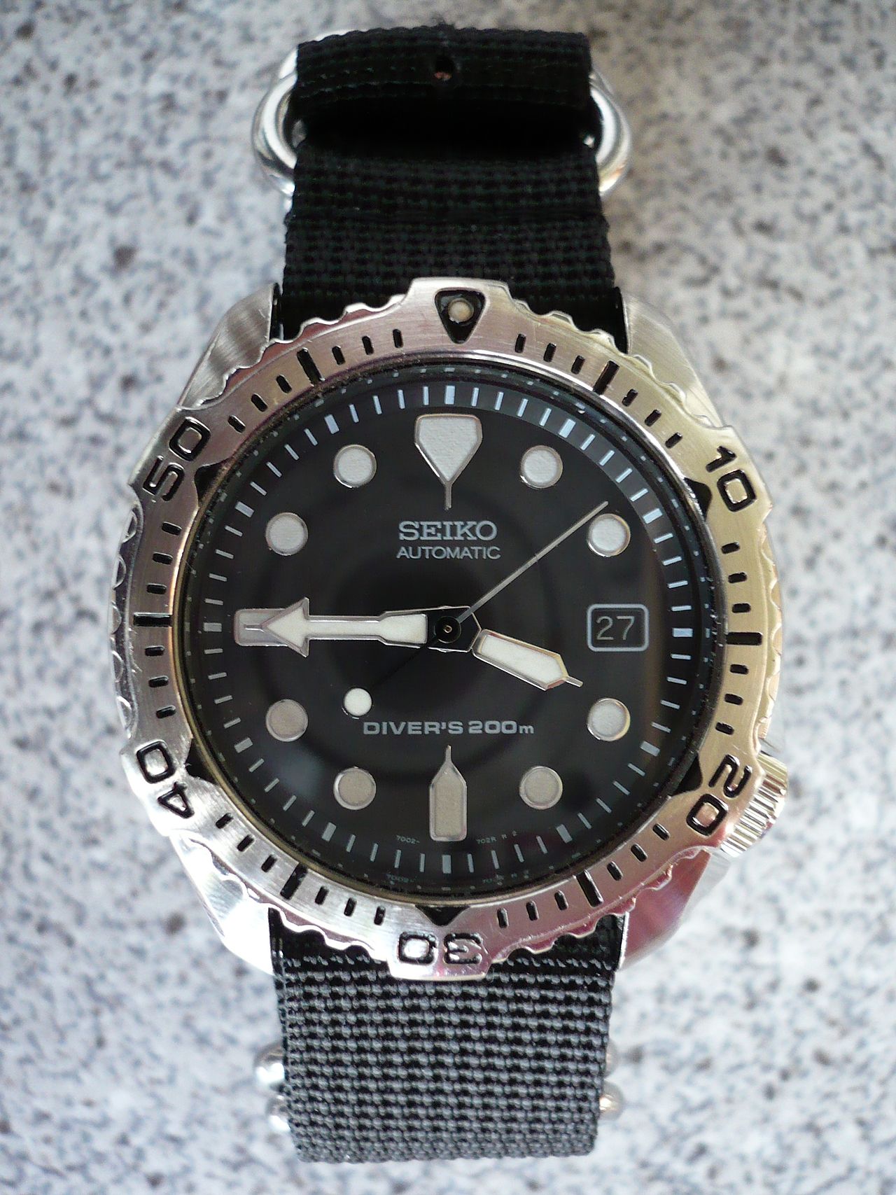 A Seiko 7002-7020 automatic diver's watch on a striped NATO strap, its black dial, dive bezel and day-date window in view, resting on a stone surface.