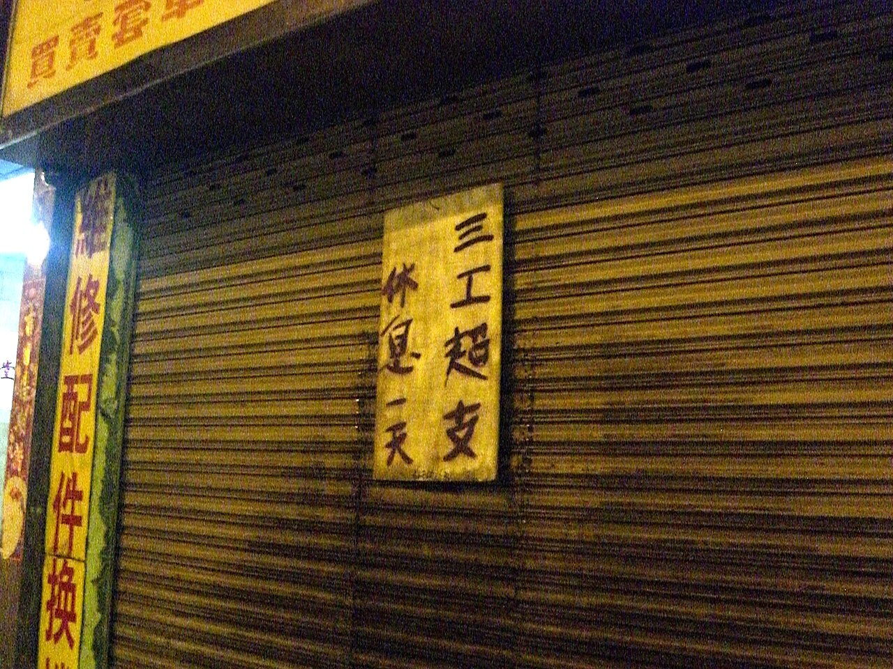 A small shop unit with its metal roller shutter pulled down, closing notices pasted across the front and a sign board still mounted above.
