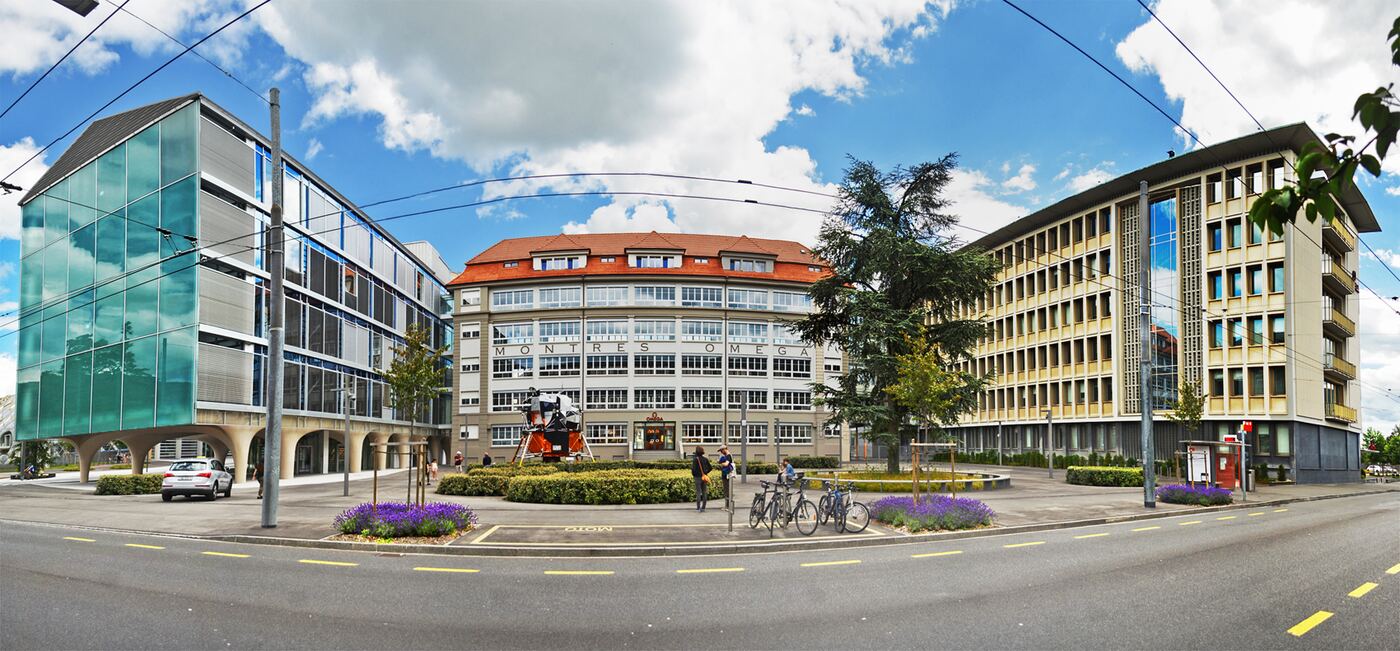 A wide view of the Swatch Group's campus in Biel, Switzerland, on a bright day with high cloud. In the centre stands a curved early-twentieth-century building with a red tiled roof and the words MONTRES OMEGA set in tall letters across its facade; a replica of an Apollo lunar module stands on the traffic island in front of it. Modern Swatch Group buildings flank it on both sides — glass and green panels to the left, a louvred stone-coloured block to the right. Tram wires cross the sky and bicycles are parked at the kerb.