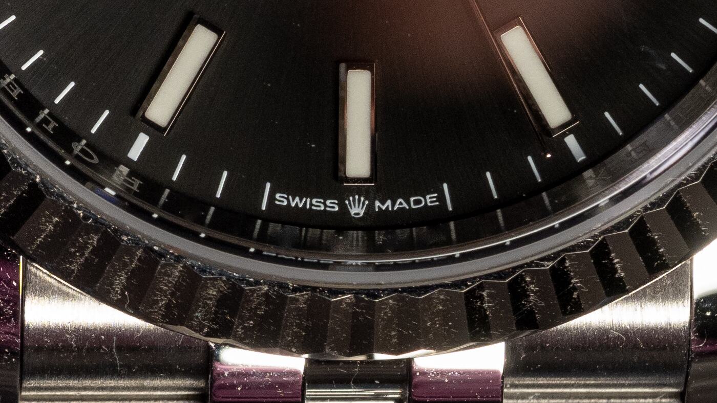 A macro photograph of the lower part of a watch dial. Across the bottom, between two short vertical bars, the words SWISS MADE are printed in small capitals, with a tiny crown between them. Above the words run the minute track and three applied baton markers; below, the fluted edge of the bezel and the links of a steel bracelet fall away out of focus.