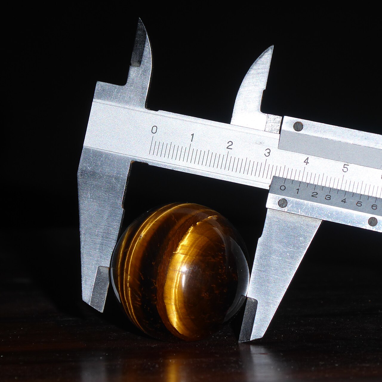 A vernier caliper closed around a polished tiger's-eye sphere against a black ground, the jaws seated on the widest point and the scale legible.