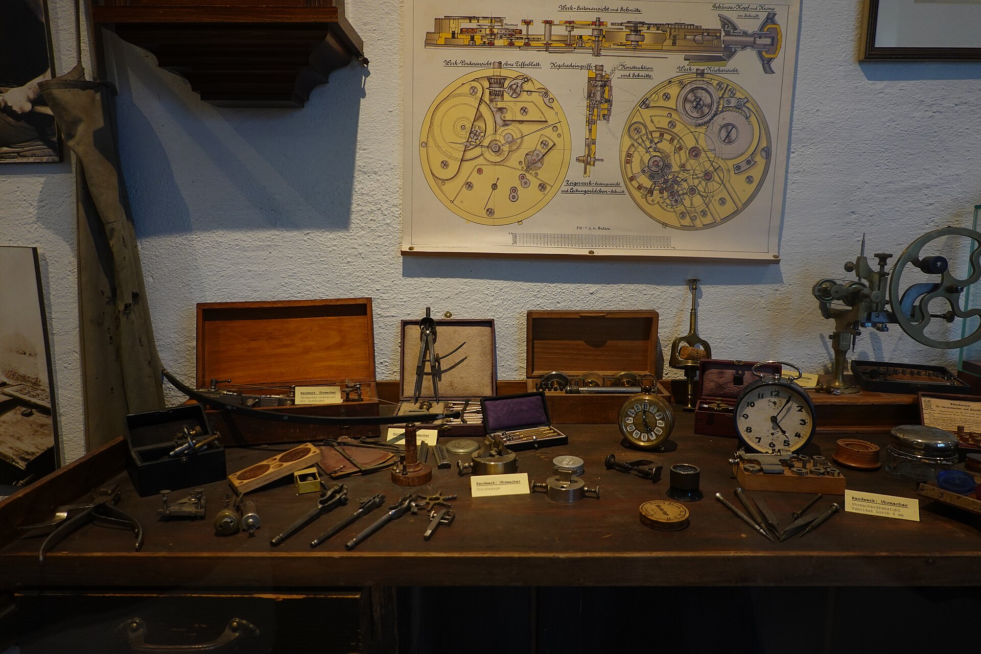 A watchmaker's bench, photographed straight on. Across the worn wooden top lie dividers, a bow-driven lathe, staking punches, hand-removing levers, movement holders, a loose-cased pocket watch and a plain alarm clock, with two open fitted boxes behind them. A large printed cutaway diagram of a pocket-watch movement hangs on the wall above. No brand name is legible anywhere in the frame.